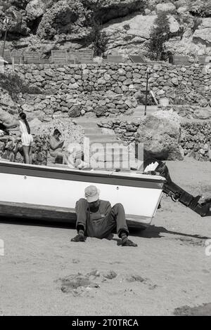 Uomo anziano con cappello appoggiato su una barca mentre dorme sulla sabbia bianca e nera della spiaggia di St Paul's Bay Foto Stock