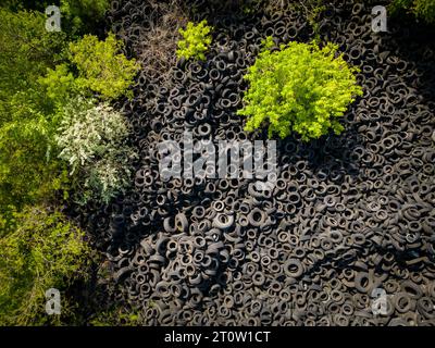 Vista aerea dall'alto del netto contrasto tra un'enorme discarica di pneumatici usati e gli alberi verdi circostanti. La scala di scarico degli pneumatici è stri Foto Stock