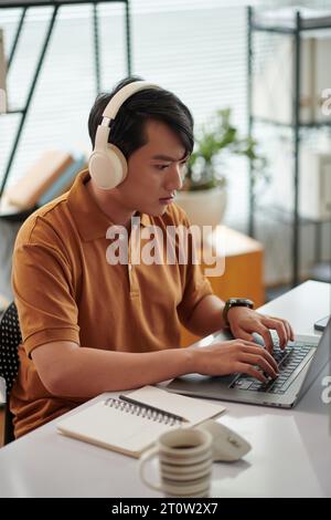 Giovane tester vietnamita che ascolta musica mentre lavora alla scrivania in ufficio Foto Stock