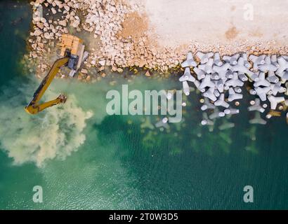 Un escavatore costruisce diligentemente un molo o un frangiflutti nel mare, il suo potente braccio che si estende dalla riva, creando una struttura resiliente in mezzo Foto Stock