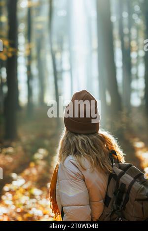 Donna con cappello in maglia e zaino che cammina nella foresta autunnale con il sole che splende tra gli alberi Foto Stock