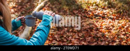 Escursionista che versa bevande calde dal thermos nella tazza da viaggio nella foresta autunnale. Ristoro durante le escursioni nella natura. Vista panoramica Foto Stock