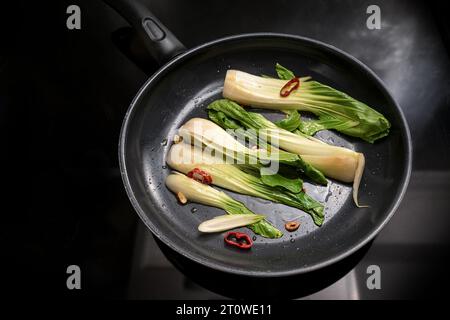 pak choi fresco o cavolo cinese con aglio, peperoncino rosso ed erbe aromatiche in padella su un piano cottura nero, cucinando un pasto vegetariano con verdure Foto Stock