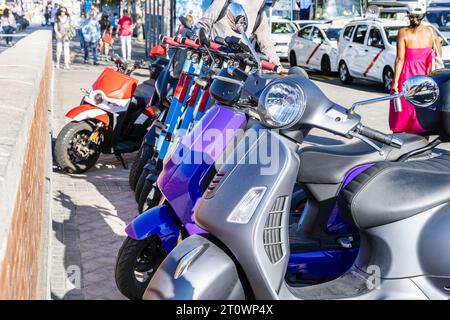 Diverse motociclette e ciclomotori di tutti i tipi e una fila di scooter elettrici blu in piedi su un marciapiede in una città europea Foto Stock