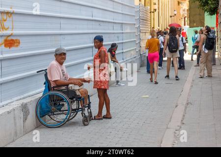 Un uomo con disabilità siede su una sedia a rotelle su un viale pedonale a l'Avana Vecchia. Una donna sta parlando con lui. Altre persone reali si stanno muovendo Foto Stock