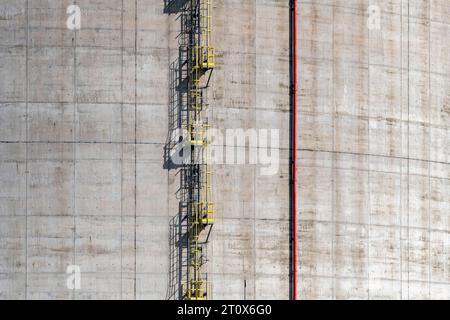 Vista di riempimento immagine di calcestruzzo con superficie liscia sul lato di un serbatoio industriale con scala verticale gialla con gabbia di sicurezza e tubazioni verticali rosse per W. Foto Stock