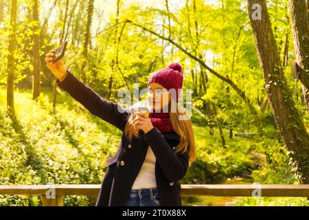 Giovane uomo con giacca nera, sciarpa e cappello di lana rosso che si diverte in un parco autunnale, scattando un selfie con la giostrina Foto Stock