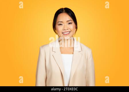 Gioiosa donna millenaria asiatica sorridente calorosamente, in posa su uno sfondo da studio arancione brillante Foto Stock