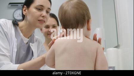 Il bambino si siede sul cambio di tavolo su appuntamento con il pediatra nel reparto ospedaliero leggero. Madre tiene il suo bambino. Medico femminile utilizza stetoscopio e controlla le condizioni del bambino. Medico al lavoro in clinica. Foto Stock