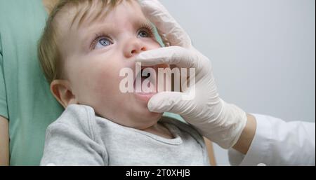 Il bambino siede tra le braccia della madre e piange. Medico femminile cerca di controllare i denti del bambino in luminoso reparto ospedaliero. Donna con bambino su appuntamento con pediatra. Medico al lavoro in clinica. Primo piano. Foto Stock