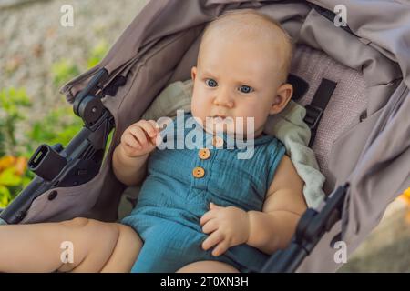 Un bambino carino, vestito con morbidi abiti in mussola, si siede tranquillamente in un passeggino, crogiolandosi in comfort e intimità in una giornata di sole Foto Stock