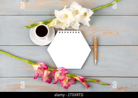 Composizione con blocco note e penna aperti, tazza di caffè nero, fiore di gladiolo bianco e rosa su tavolo di legno blu. Conce desktop per ufficio festivo Foto Stock