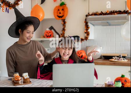 Una giovane ragazza asiatica felice e felice in costume di Halloween ama guardare i cartoni animati di Halloween su un portatile con la mamma in cucina, per festeggiare Foto Stock