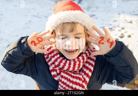 Un bambino sorridente e allegro con il cappello di Babbo Natale e la sciarpa rossa a righe chiudono gli occhi e fanno desiderare. i numeri del nuovo anno 2024 sono scritti su palme. il ragazzo è cameriere Foto Stock