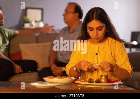 Giovane donna che organizza puja thali in occasione di Raksha Bandhan mentre suo padre e suo fratello sono seduti dietro Foto Stock