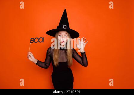 Ritratto fotografico di una bella adolescente bionda che tiene il segno del boo Scratch vestito a mano nero vestito da halloween isolato su sfondo di colore arancione Foto Stock