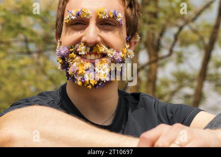 Circondato da alberi in parchi, boschi e foreste, un giovane uomo ha fiori primaverili che si sostituiscono con i capelli del viso. Seduto in mezzo alla natura, abbracciando le ginocchia Foto Stock