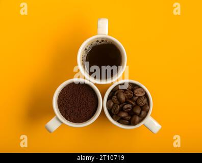 Vista dall'alto di tre tazze di caffè bianche su sfondo giallo. Uno con caffè macinato, l'altro con chicchi di caffè e l'altro con caffè liquido. Foto Stock