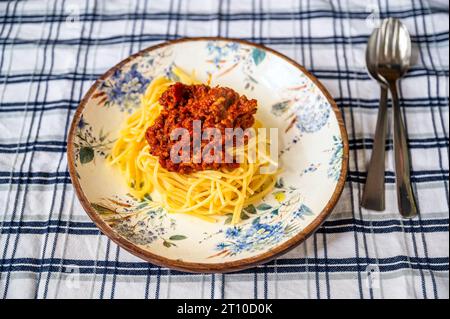 Spaghetti con carne macinata e salsa di pomodoro (spaghetti bolognesi) in piatto decorativo, forchetta e cucchiaio su tovaglia a scacchi. Foto Stock