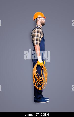 Scatto integrale, vista laterale ritratto a metà faccia di un elettricista seducente e tranquillo con setole in tuta, camicia, elmetto con cavo arrotolato Foto Stock