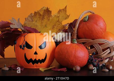 Buon Halloween. Secchio di zucca in plastica con un sorriso malvagio, zucche mature, cesto e foglie autunnali. Composizione per Halloween su sfondo arancione Foto Stock