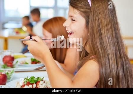 Vista laterale di Happy Girl che mangia con forchetta durante il pranzo nella caffetteria scolastica Foto Stock
