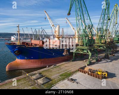 Una nave portarinfuse caricata o scaricata con grano è ancorata in un porto affollato, con lavoratori e macchinari visti sullo sfondo. Foto Stock