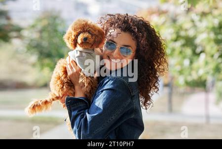 Ritratto di un piccolo cane dai capelli rossi, mini barboncino con donna riccia guarda la macchina fotografica e sorride a Park. PET sembra il proprietario. Foto Stock