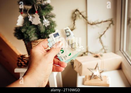 Concetto di bilancio e finanze natalizie. Mano di una donna che tiene soldi contro l'albero di Natale e gli ornamenti in casa. Foto Stock