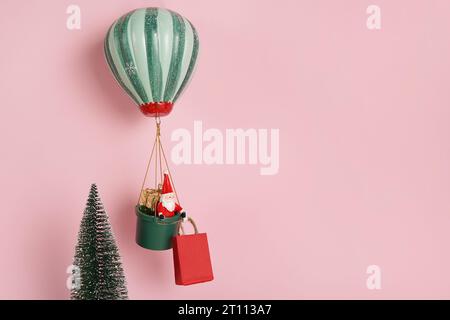 Biglietto per l'anno nuovo. Il giocattolo Babbo Natale vola sulla mongolfiera vicino all'albero di Natale e tiene in mano un sacchetto di carta rossa. sfondo rosa con spazio di copia Foto Stock