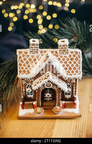 Una piccola casa di pan di zenzero con glassa in piedi sul tavolo. Soggiorno con luci e albero di Natale. Atmosfera natalizia Foto Stock