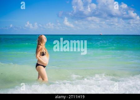 Radiosa donna incinta in costume da bagno, sullo sfondo mozzafiato di un mare turchese. Bellezza serena della maternità vicino alla riva Foto Stock