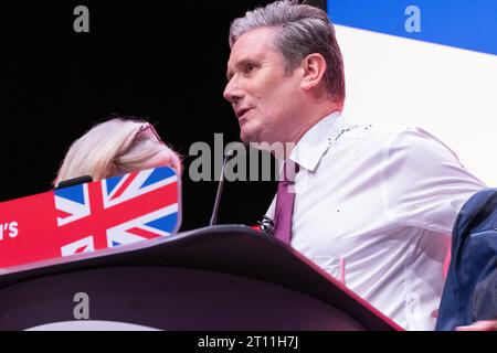 Keir Starmer rimuove la giacca dopo che il manifestante copre Keir Starmer con i glitter durante l'inizio del discorso dei leader. Il manifestante è stato lottato a terra e rimosso dalla sicurezza. Sir Keir si spazzolò i capelli si tolse la giacca e proseguì col discorso. La sua camicia, le mani e il pavimento intorno a lui erano ricoperti di glitter e sono rimasti così per tutto il discorso. Conferenza del lavoro 2023. Immagine: Garyroberts/worldwidefeatures.com Foto Stock