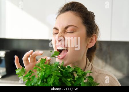 Ritratto di una donna attraente che morde prezzemolo, mangia verdure fresche ed erbe in cucina, godendo di uno stile di vita sano e di cibo Foto Stock