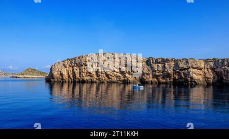 una piccola barca si avvicina alla scogliera calcarea di una delle isole kornati che si riflette nel calmo mare blu Foto Stock