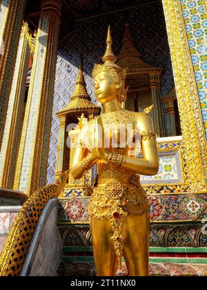 Adagiato sullo sfondo di un tempio impreziosito da piastrelle a mosaico blu e oro, una statua dorata di una donna vestita in tradizionale abito thailandese captur Foto Stock