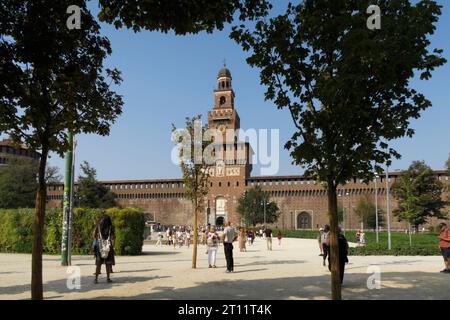 Turisti e visitatori camminano attraverso il parco verso la storica facciata in mattoni e la torre dell'orologio del Castello Sforzesco a Milano, Italia, Europa Foto Stock
