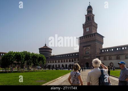Turisti e visitatori camminano attraverso il parco verso la storica facciata in mattoni e la torre dell'orologio del Castello Sforzesco a Milano, Italia, Europa Foto Stock