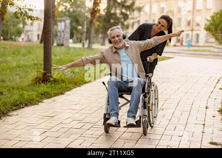 Gioioso uomo disabili maturo in sedia a rotelle indossando le cuffie divertirsi durante una passeggiata in città assistita da una bella giovane infermiera Foto Stock