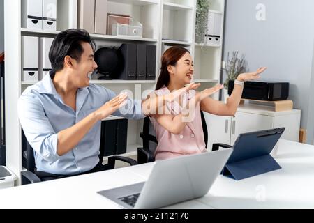 Felicissimo uomo d'affari e donna asiatica sorridendo e ballando con felicità e celebrando il successo mentre si è seduti sulla scrivania con computer portatile e tablet in ufficio. - Un posto di lavoro di successo e divertente. Foto Stock