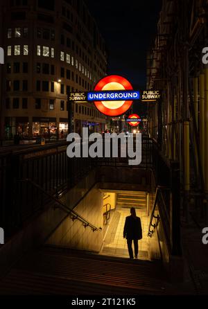 Londra, Regno Unito: Ingresso alla stazione della metropolitana Monument di notte con un cartello illuminato della metropolitana di Londra e una persona che cammina lungo i gradini. Foto Stock