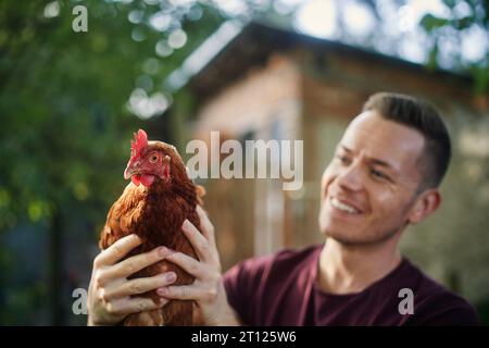 Ritratto di un contadino sorridente mentre si trova all'esterno del pollaio e tiene la gallina in un'azienda agricola biologica. Foto Stock