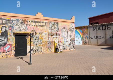 Ouarzazate, Marocco - 10 ottobre 2023: Muro dipinto con i volti di artisti e graffiti diversi e un ragazzo seduto su una scala. Foto Stock