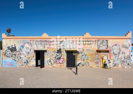 Ouarzazate, Marocco - 10 ottobre 2023: Muro dipinto con i volti di artisti e graffiti diversi e un ragazzo seduto su una scala. Foto Stock