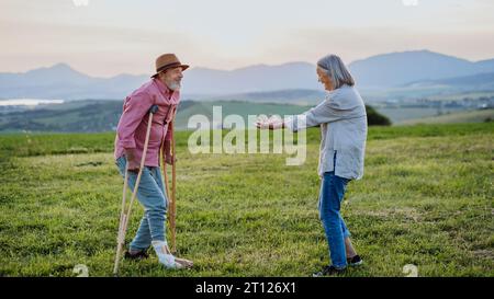 Uomo anziano con gamba rotta e stampelle. Guarigione, riabilitazione dopo lesioni o interventi chirurgici in natura. Moglie che aiuta il marito a camminare durante Easy Foto Stock