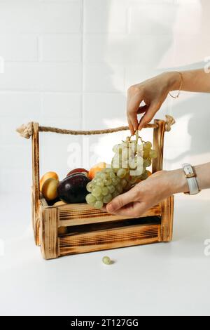 Ci sono frutta in un cesto su un tavolo bianco - prugne, albicocche e uva nelle mani dei donne. Telaio verticale Foto Stock