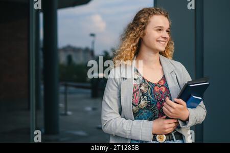 Ritratto all'aperto di uno studente allegro con libri di esercizi in piedi vicino all'edificio del college, che guarda lontano e sorride. Foto Stock