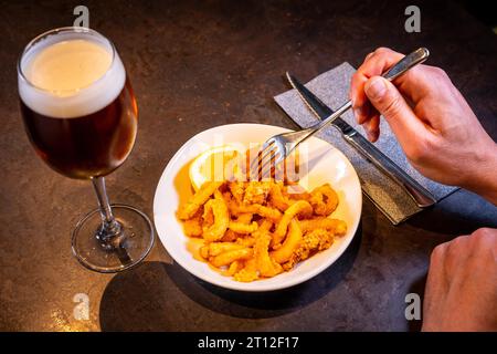 Un cliente che mangia calamari impanati con limone accompagnato da una birra su sfondo nero, su un piatto bianco Foto Stock