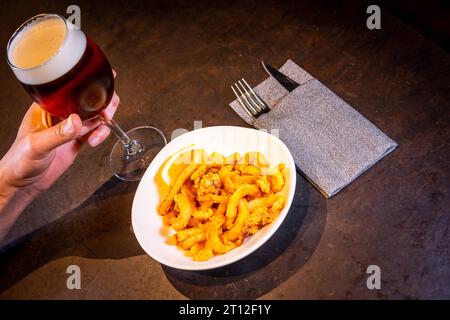 Un cliente che mangia calamari impanati con limone accompagnato da una birra su sfondo nero, su un piatto bianco Foto Stock