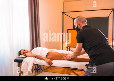 Massaggio a casa, facendo un massaggio a casa del cliente sulle gambe. Con una maschera facciale nella pandemia di coronavirus Foto Stock